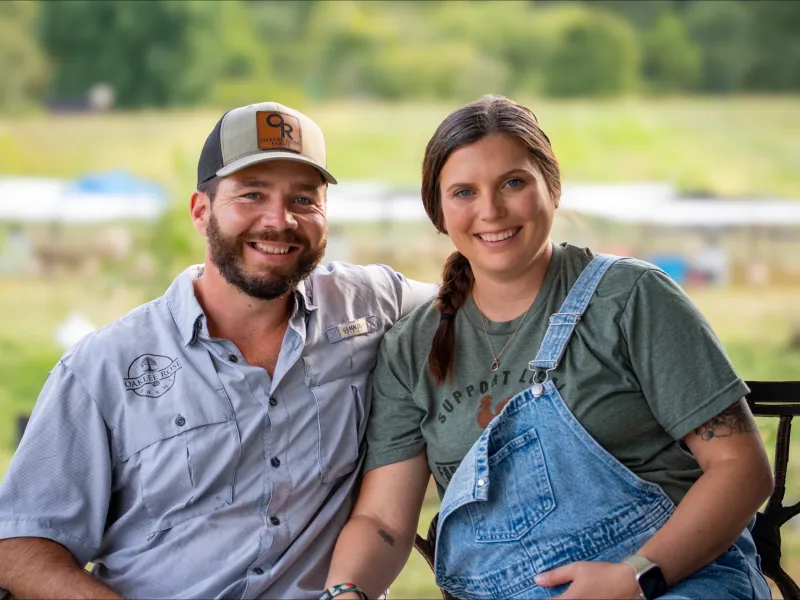 Nick and his wife sit outside with their farm in the background