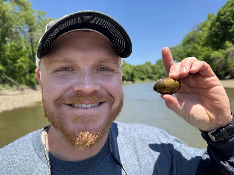 Trevor Starks holds up a mollusk in front of a river on a sunny blue day