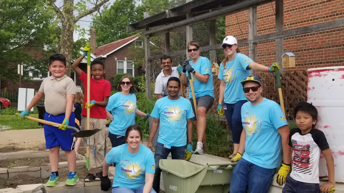 Youth and teen volunteers in blue shirts hold garden tools and learn against garden beds.