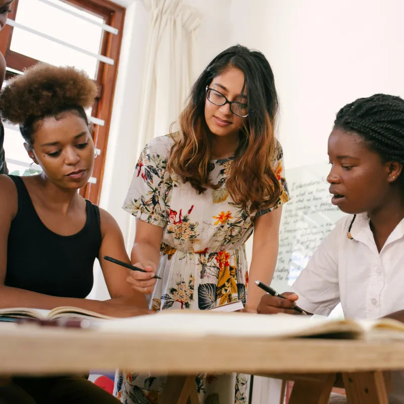 Three female presenting people of various skin colors, hair styles, and professional workplace clothes, in a bright room, appearing to collaborate on something on a table.