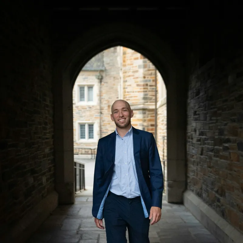 Smiling, male appearing person in casual business clothes of light blue shirt and dark blue jacket walking through a passageway between stone buildings.